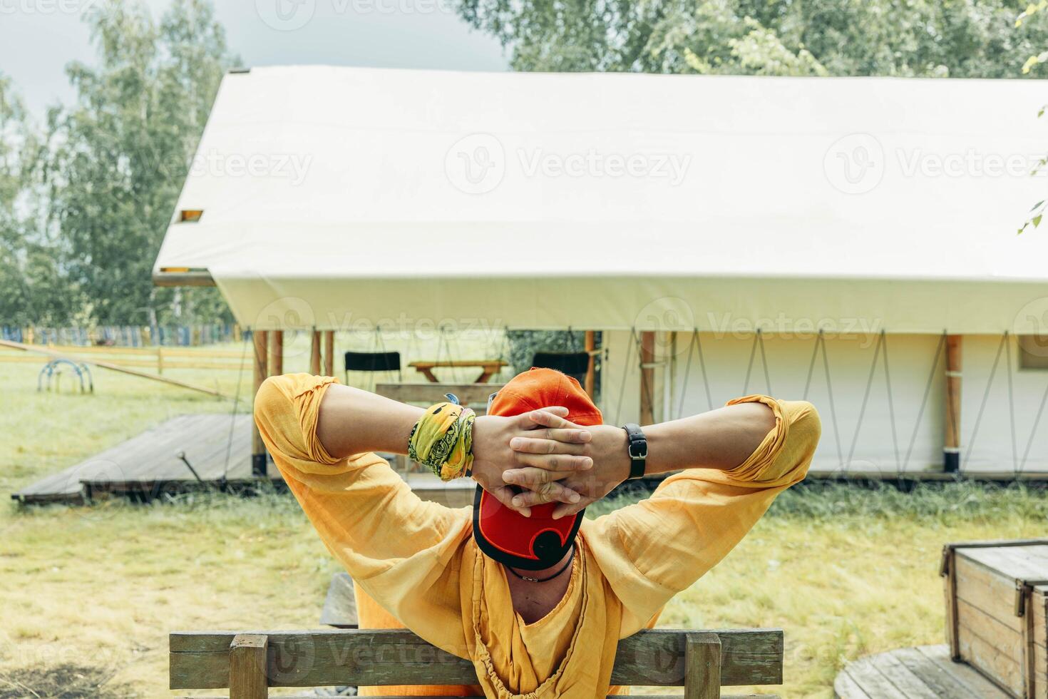 man in orange cap and yellow hoodie leaning back on wooden bench with hands behind head in front of glamping tent in forest meadow on summer day, travel eco tourism slow lifestyle retreat photo