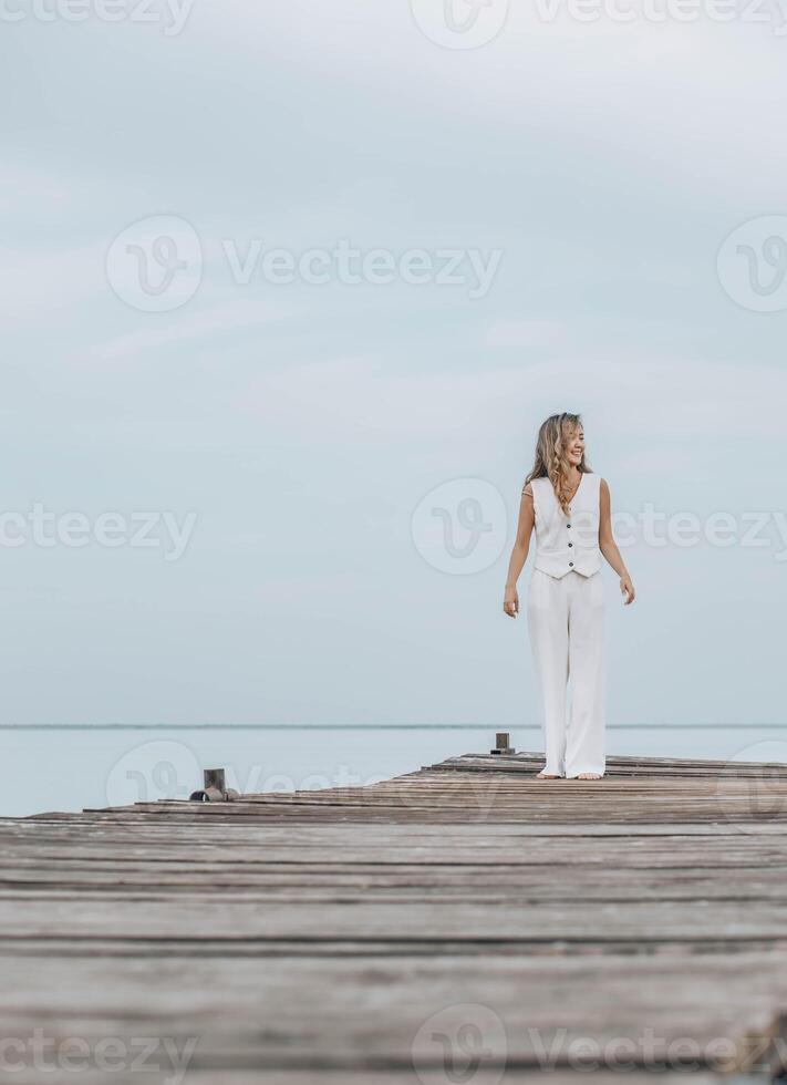 Asian woman in white outfit stands on wooden pier, calm lake, peaceful overcast, viewed from behind with contemplative mood, mindfulness, travel retreats, wellness, vertical, copy space photo