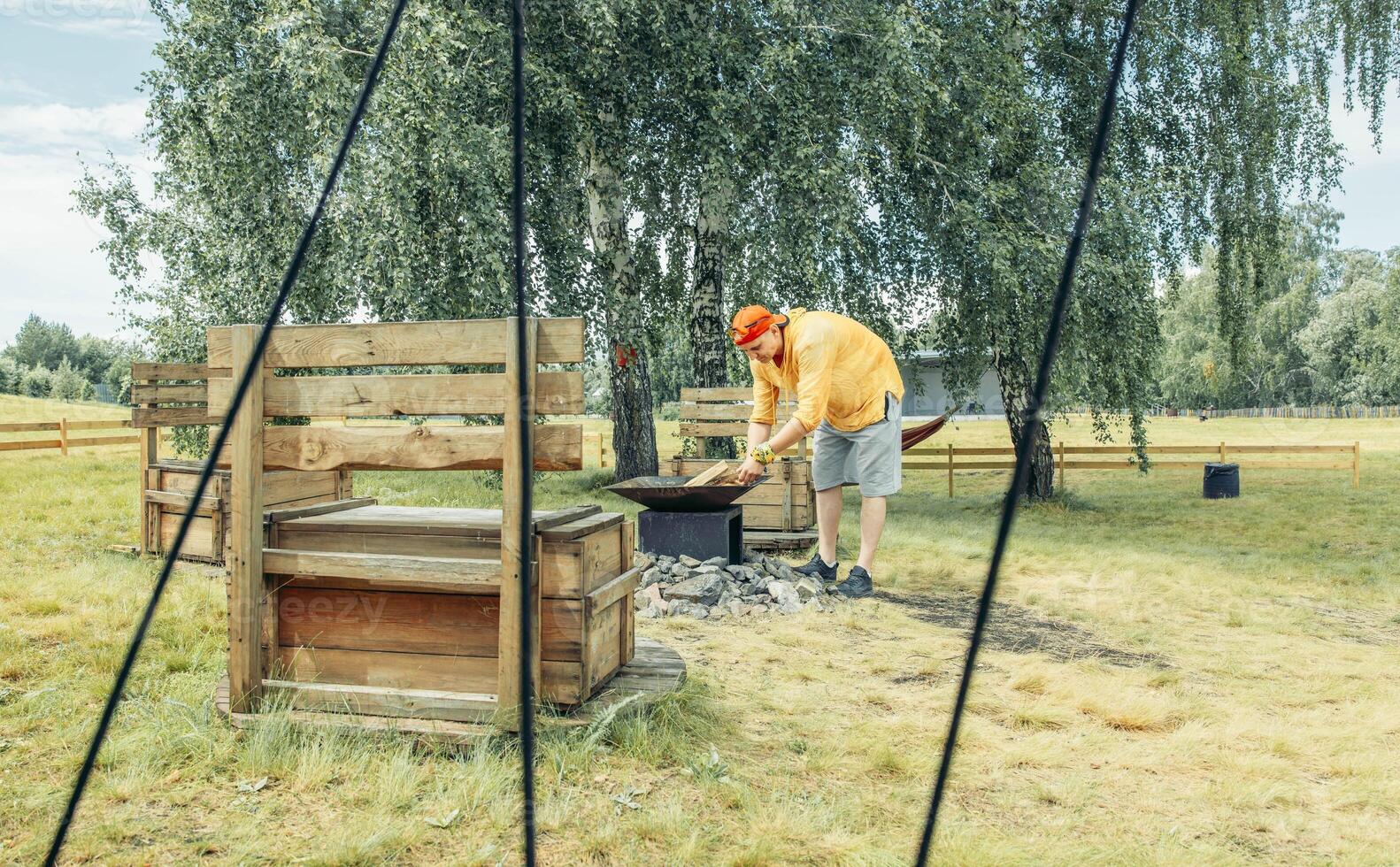 man in red cap, in yellow clothes near outdoor fire pit surrounded by stones with rustic benches seen through tent ropes, camping equipment, travel agency, outdoor adventure photo