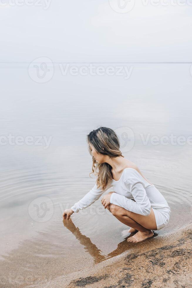 unrecognizable woman squatting barefoot at lake edge creating water ripples with fingers, gazing at water with serene expression, calm horizon under cloudy sky, mindfulness, wellness, retreat photo
