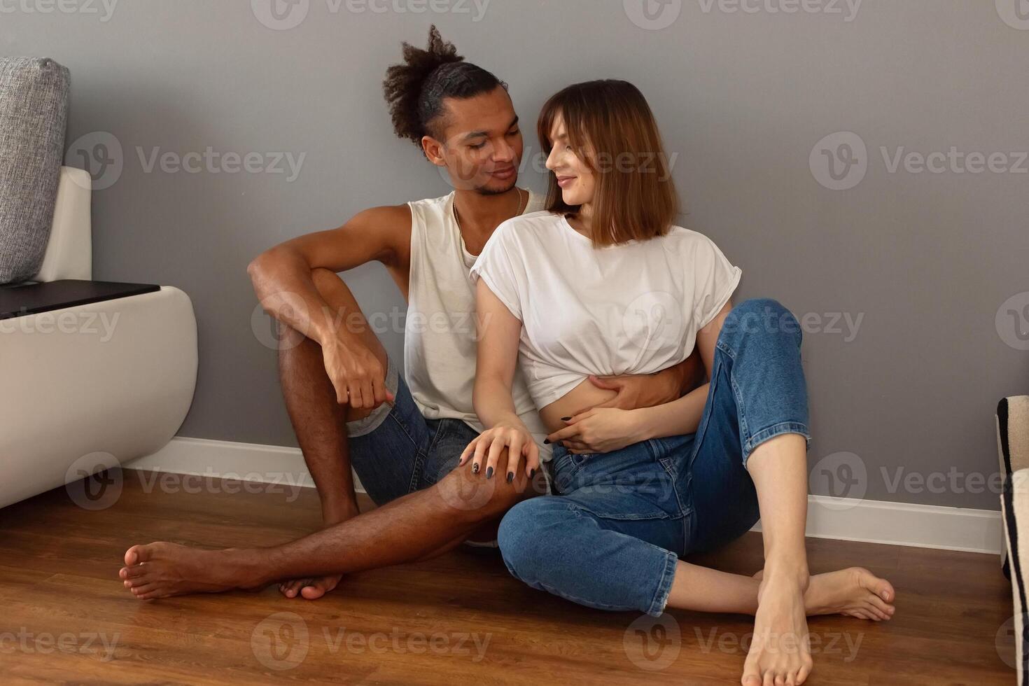 A couple are sitting at home on the floor against a gray wall photo