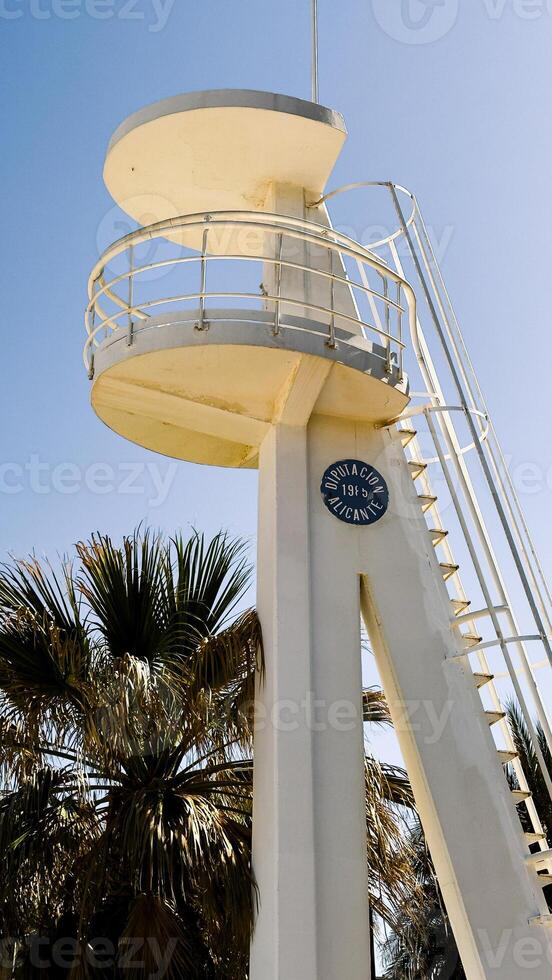 White lifeguard tower stands near palm trees in Alicante, Spain. Image suitable for beach safety, tourism, or coastal architecture context photo