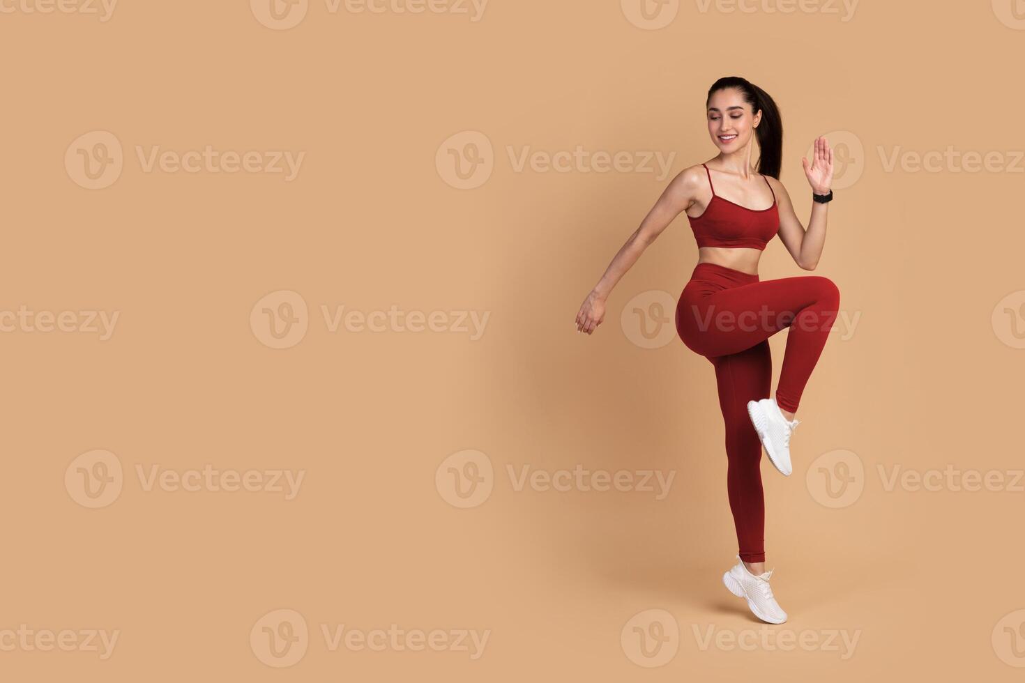 Warmup. Full body length shot of smiling young fitness model in sportswear standing on one leg and touching knee with her elbow, doing high knees exercise isolated over pastel studio background photo