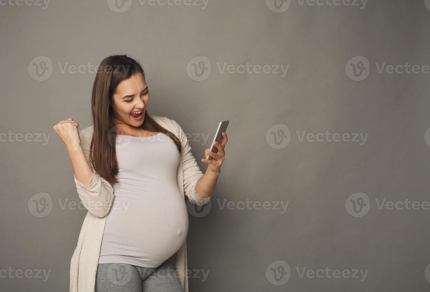 Surprised pregnant woman using smartphone copy space. Excited expectant lady with open mouth looking at mobile phone, gray studio background. Success, modern technologies, pregnancy concept photo