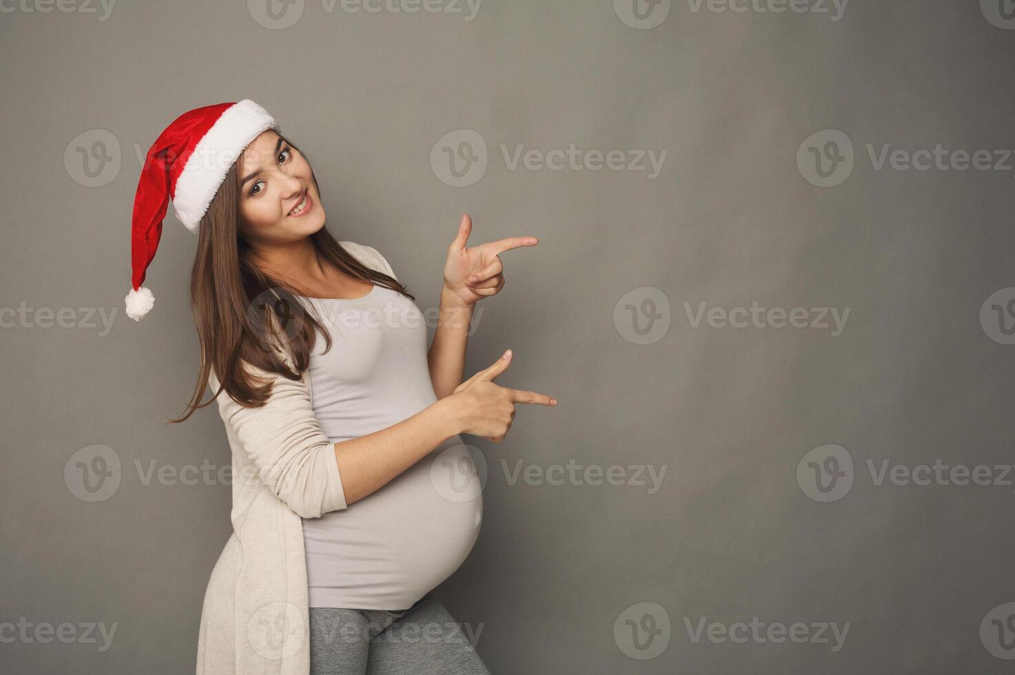 Young beautiful pregnant woman in Santa hat pointing aside portrait. Cheerful expectant girl showing something, gray studio background, copy space. Christmas sale, pregnancy concept photo