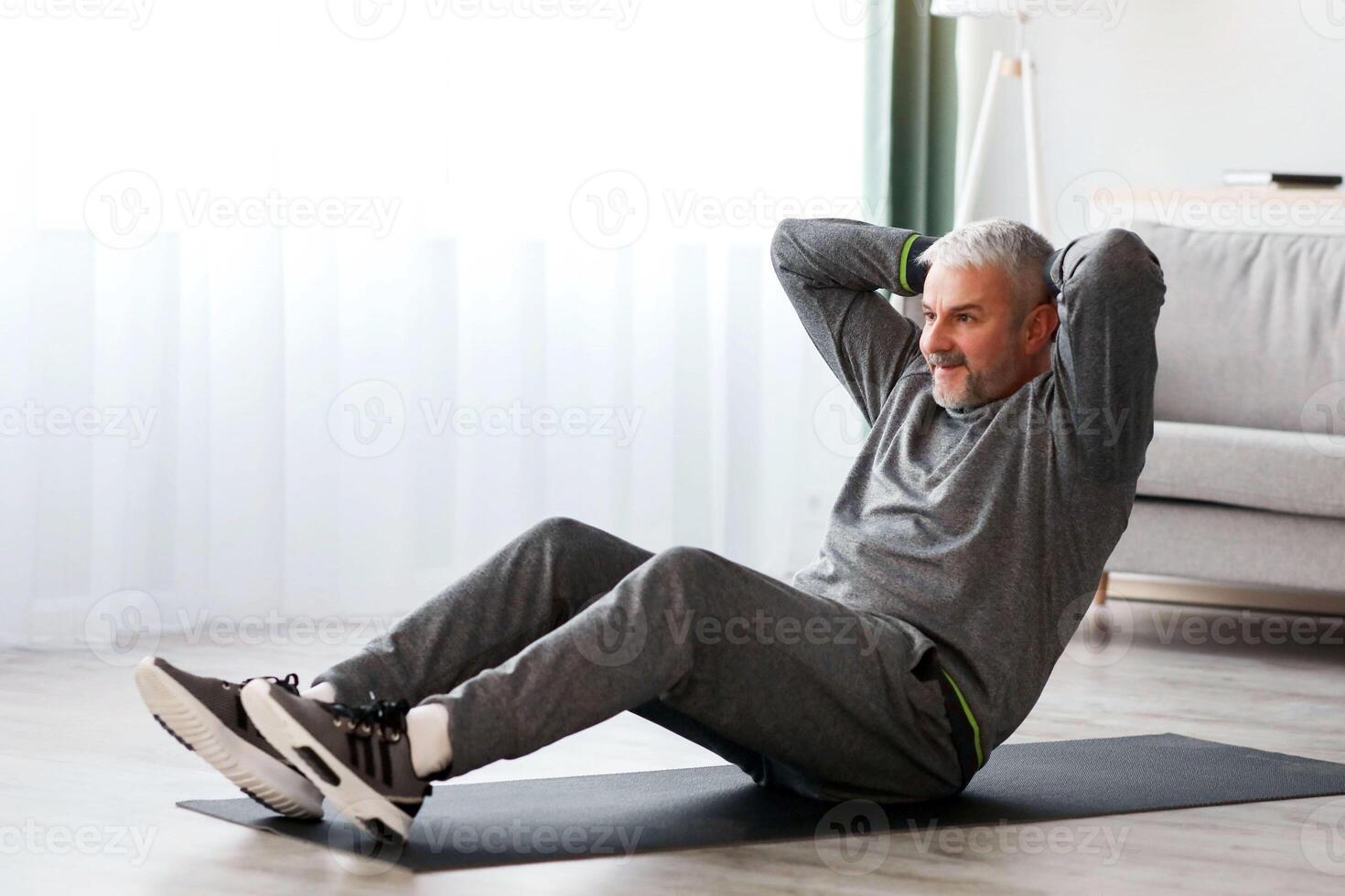 Athletic bearded senior man in grey sportswear doing exercises for abs at home, sitting on fitness mat and crunching, copy space. Healthy lifestyle, sport on self-isolation for older people photo