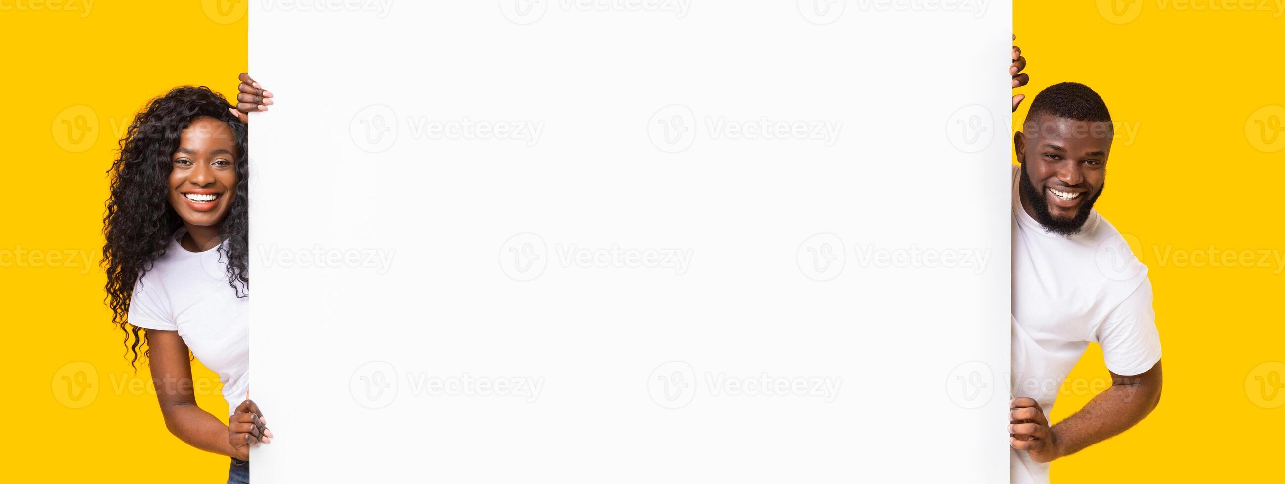 Happy black man and woman standing next to blank placard for advert. Cheerful african american couple in love holding empty board for advertisement or text over yellow studio background, panorama photo