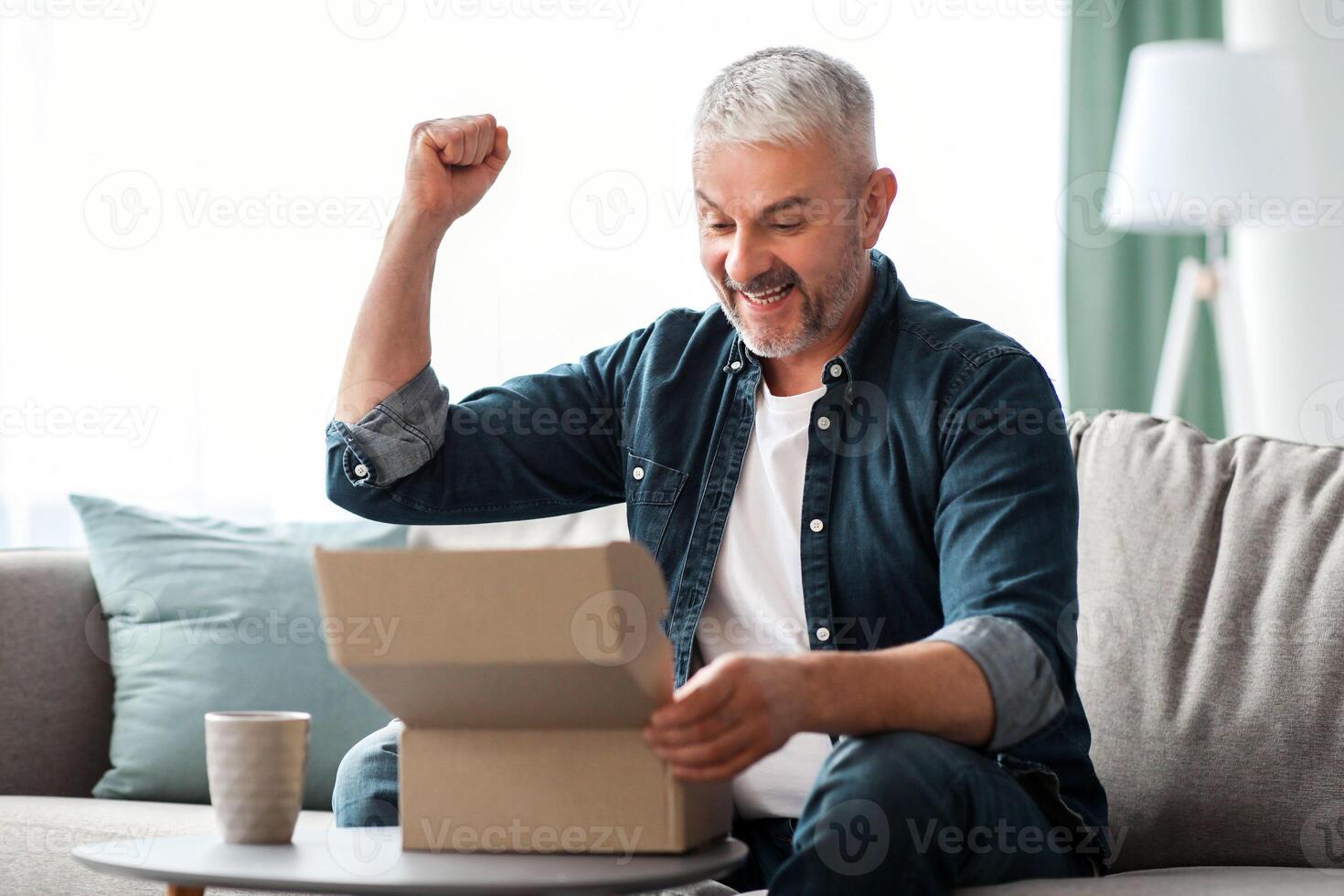 Happy senior man customer checking mail box, raising fists up and screaming, sitting on sofa at home, copy space. Emotional elderly man opening delivery, checking order. Delivery concept photo