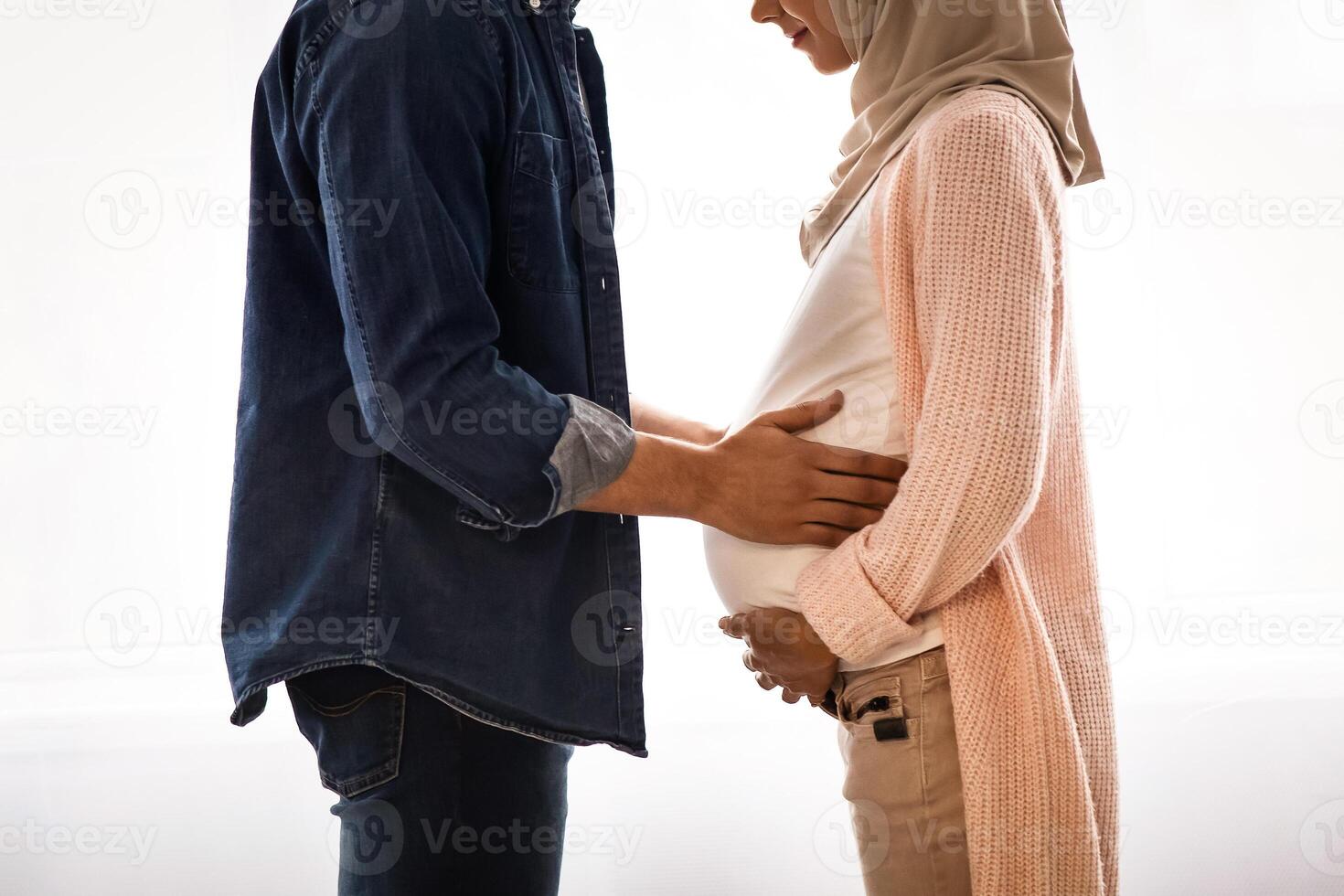 Young religious muslim couple expecting baby, standing together near window at home, caring millennial arab husband tenderly touching belly of his pregnant wife in hijab, side view with copy space photo