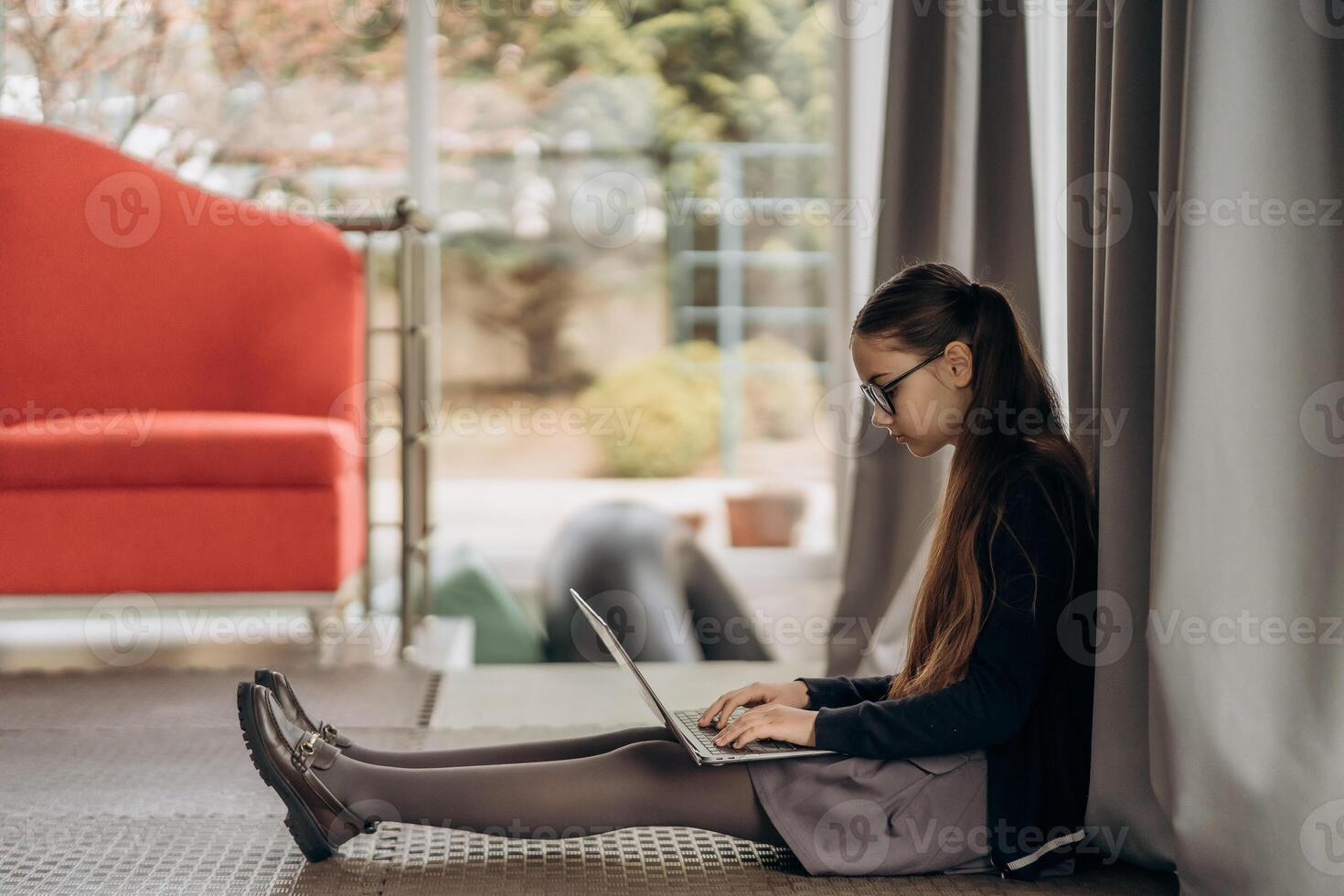 Teen girl in glasses working on laptop while sitting on floor near window with natural light soft curtains and minimalist background showing calm indoor lifestyle focused digital routine and remote photo