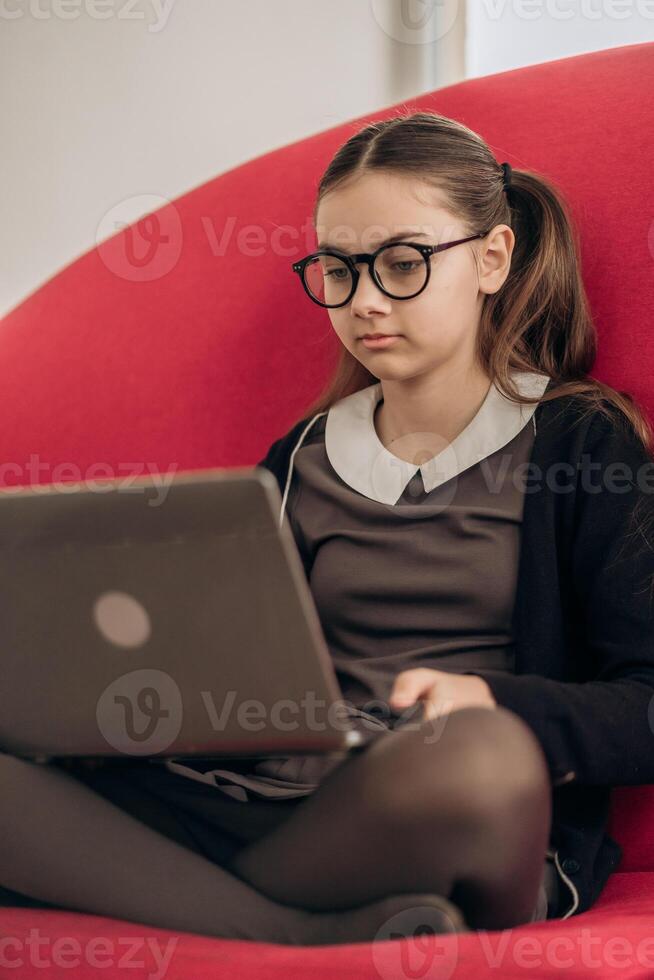 Focused girl in school uniform sitting with laptop on red sofa wearing glasses deeply concentrated on screen representing young generation adapting to digital education hybrid homeschool Gen Alpha photo