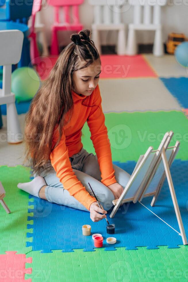 Girl painting at wooden easel on colorful foam mat using brush and jars of paint in creative classroom showing independence imagination and peaceful concentration during early childhood artistic photo