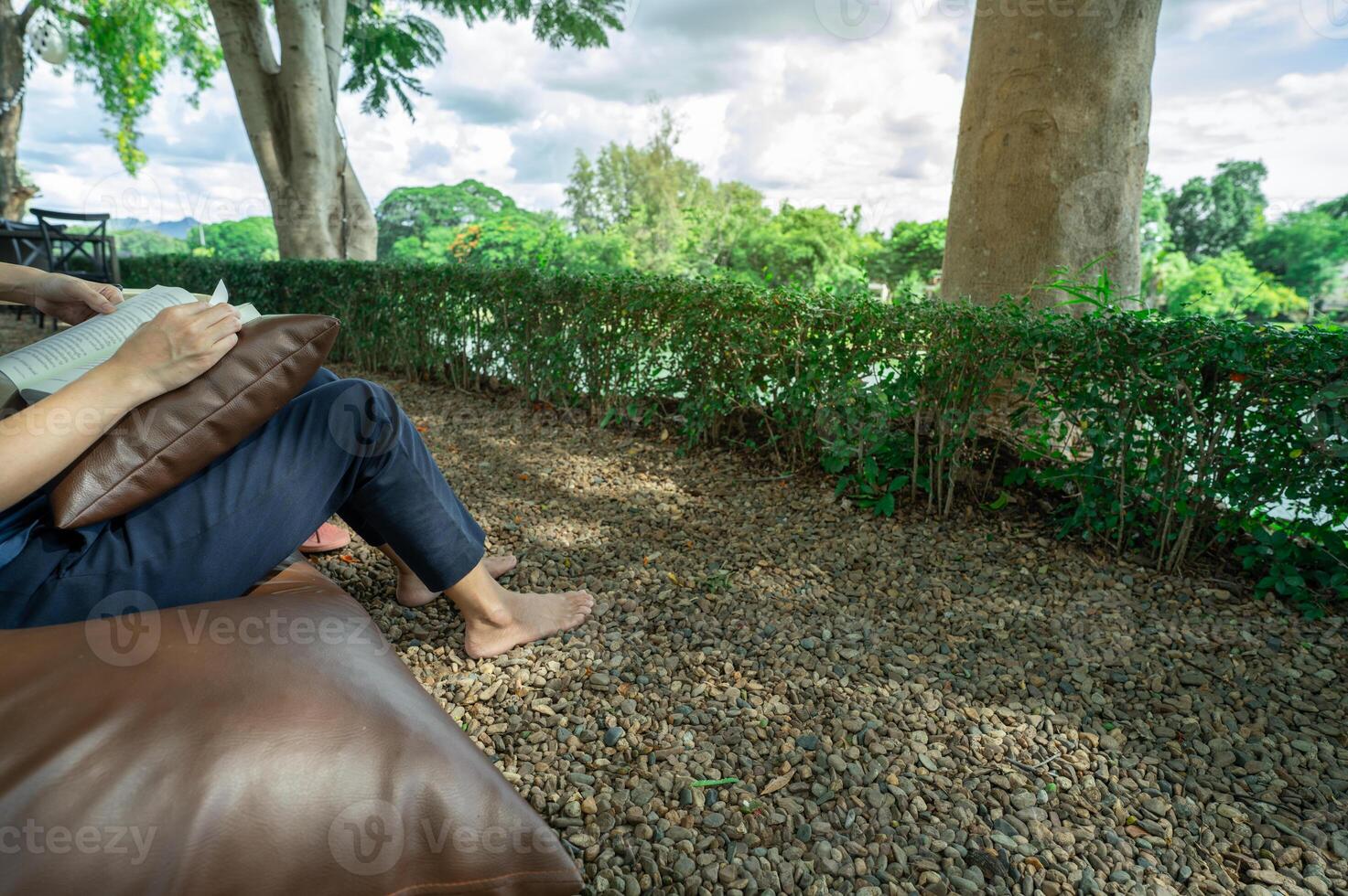 Barefoot woman enjoying digital detox by reading book outdoors at riverside hotel resort with natural stone ground and cool breeze for peaceful self-reflection and mental wellness escape. Self-care. photo