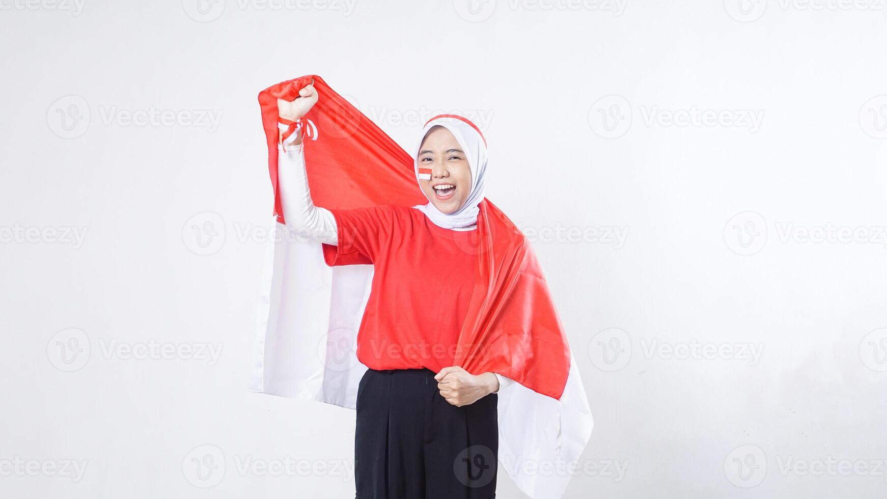 Excited Asian girl celebrate Indonesia independence day in red and white outfit showing victory pose with Indonesian flag draped around her back like a cape isolated on white background photo