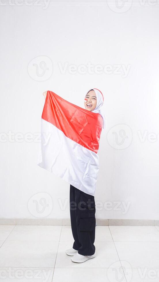 Happy Asian girl celebrate Indonesia independence day in red and white outfit holding and wrapping herself with Indonesian flag isolated on white background photo
