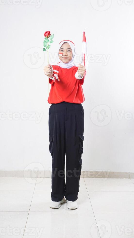 Beautiful Asian girl celebrate Indonesia independence day in red and white outfit giving a rose and tiny Indonesia flag isolated on white background photo