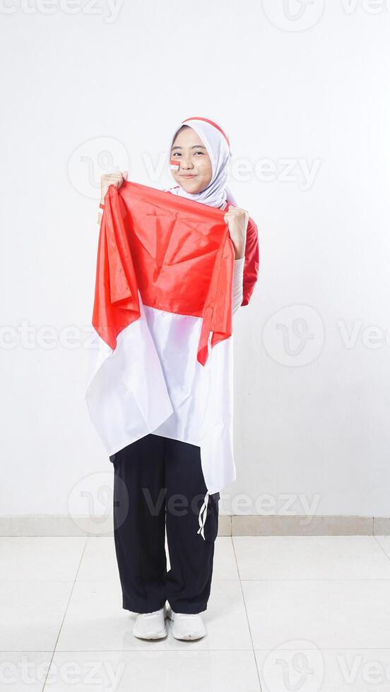 Happy Asian girl celebrate Indonesia independence day in red and white outfit holding Indonesian flag with two hands isolated on white background photo
