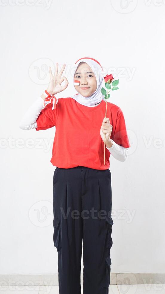 Beautiful Asian girl celebrate Indonesia independence day in red and white outfit showing OK sign hand gesture while holding a rose isolated on white background photo