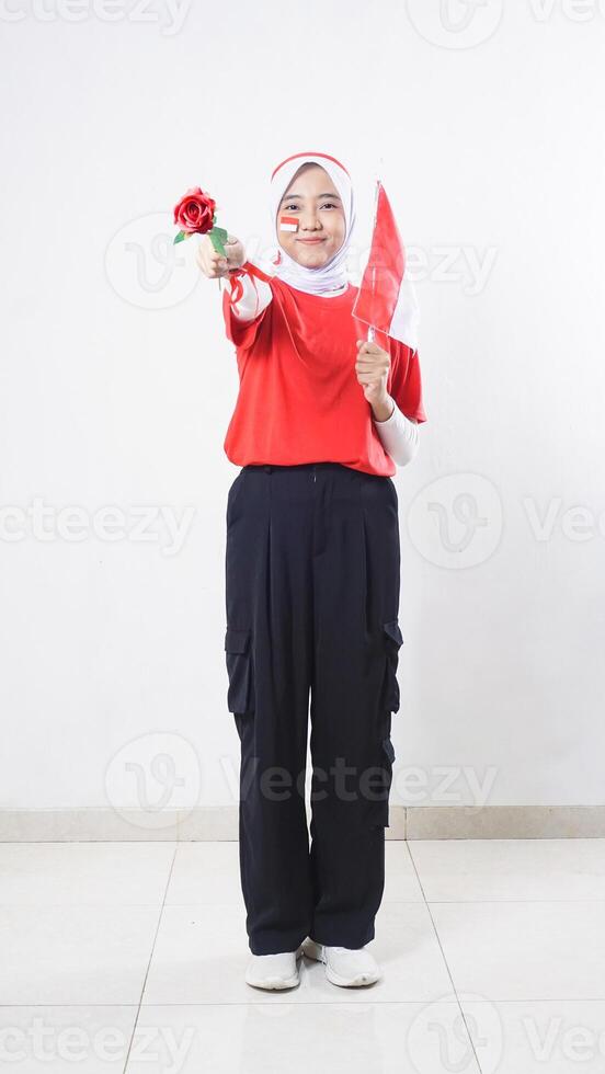 Beautiful Asian girl celebrate Indonesia independence day in red and white outfit giving a rose and holding tiny Indonesia flag isolated on white background photo