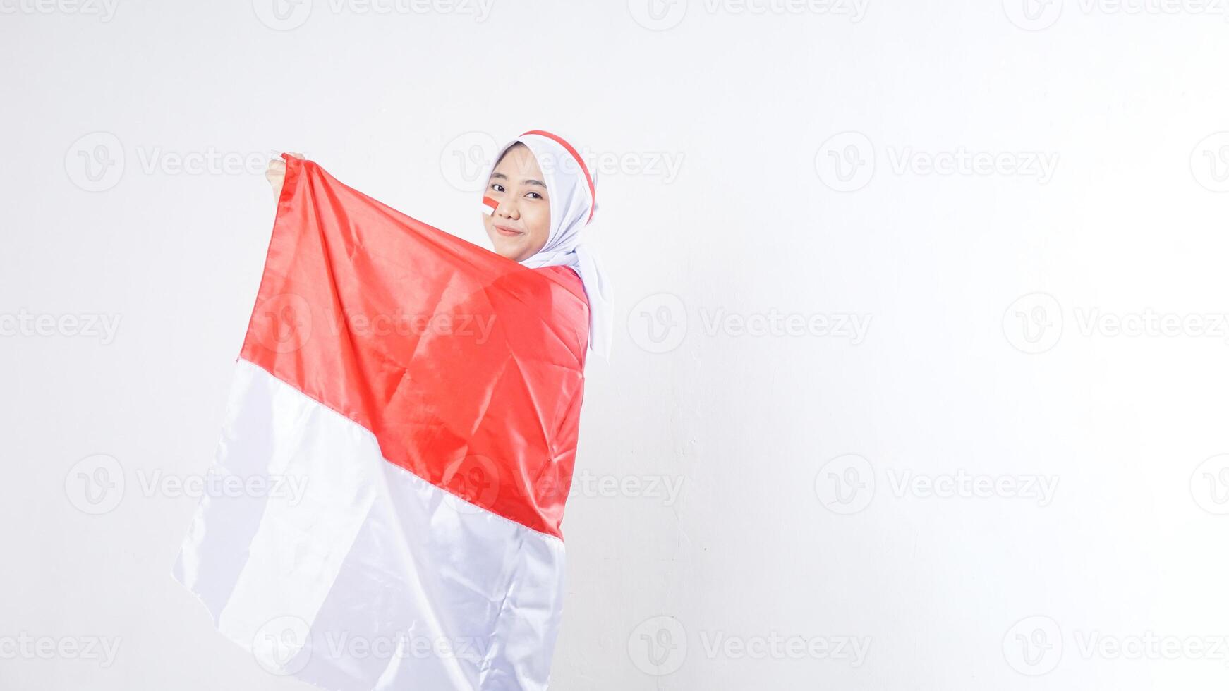 Beautiful Asian girl celebrate Indonesia independence day in red and white outfit holding and wrapping herself with Indonesian flag isolated on white background photo