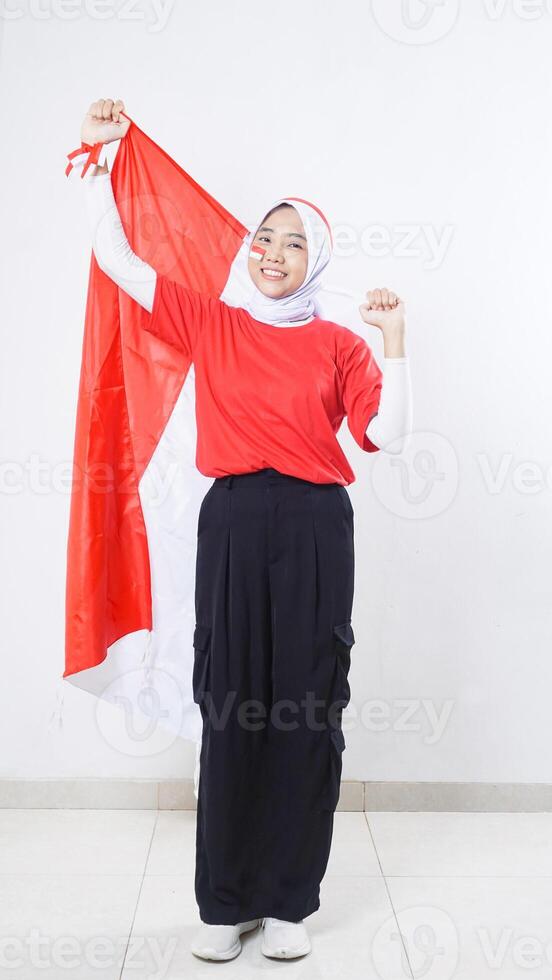 Happy Asian girl celebrate Indonesia independence day in red and white outfit holding Indonesian flag behind her back isolated on white background photo