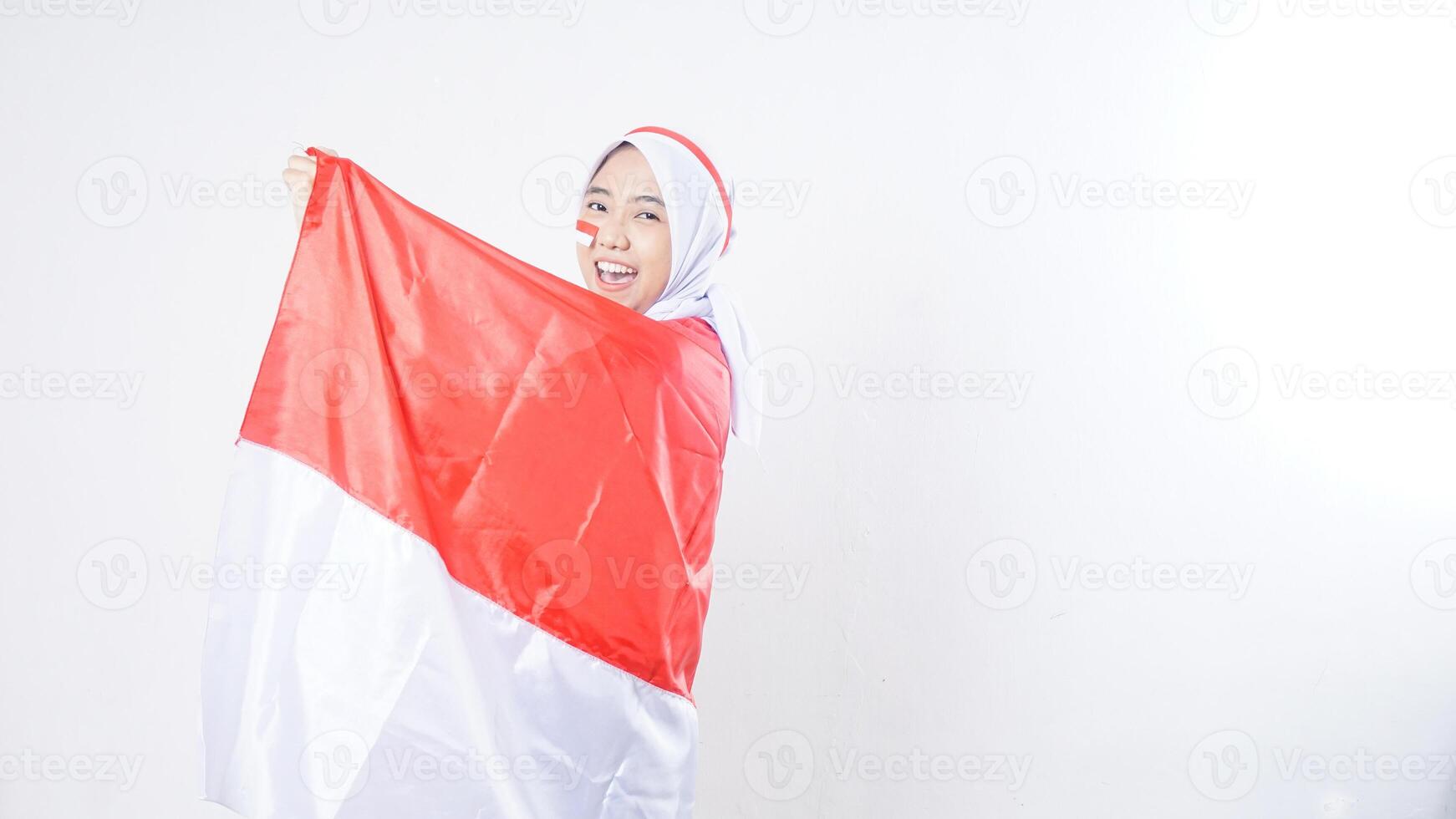 Excited Asian girl celebrate Indonesia independence day in red and white outfit holding and wrapping herself with Indonesian flag isolated on white background photo