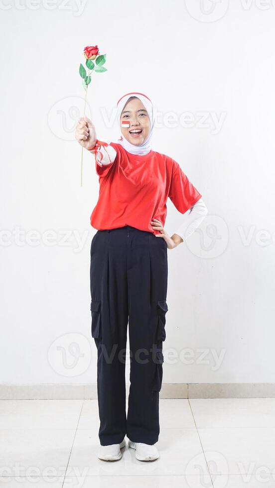 Excited Asian girl celebrate Indonesia independence day in red and white outfit giving a rose isolated on white background photo