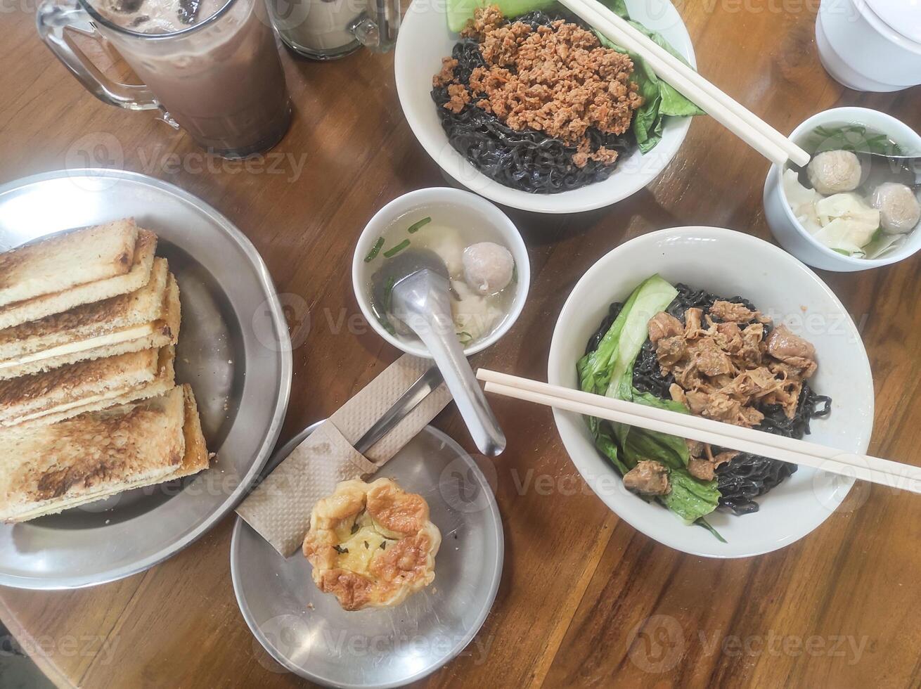 Tabletop shot of a typical Indonesian lunch menu. The dish consists of chicken noodles with meatball soup, a cold chocolate drink, and desserts of toast and egg tart. Tasty Indonesian gourmet photo