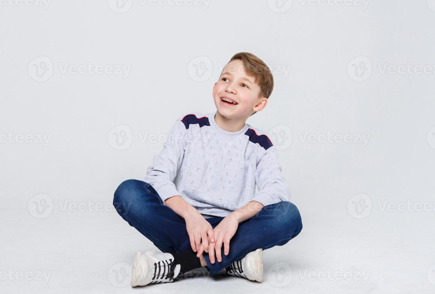 Portrait of happy smiling boy in braces sitting on the floor at white studio background. Kid in casual clothes looking up, copy space photo