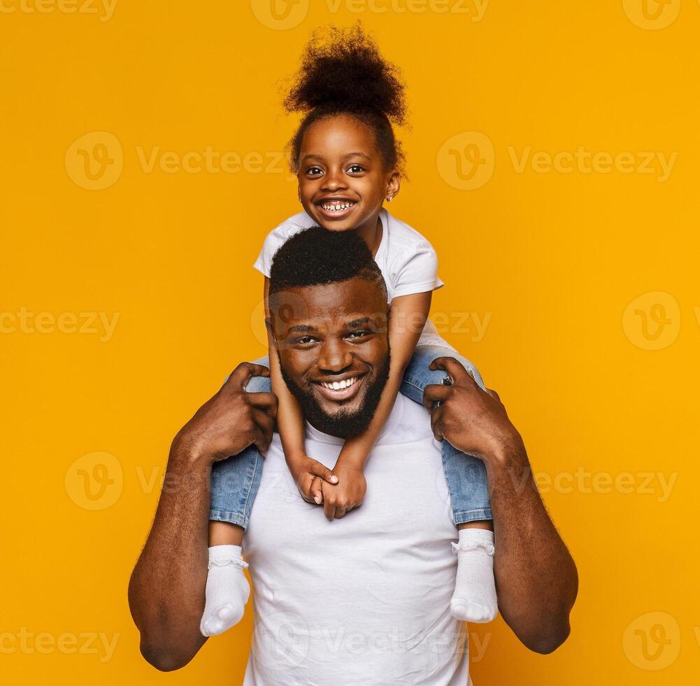 Cheerful portrait of black dad and little daughter sitting on father shoulders over yellow background photo