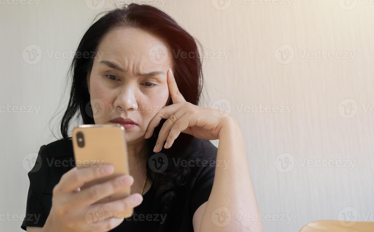Depressed mature Asian woman sit and looking on phone, lost in thoughts, thinking about problem. photo