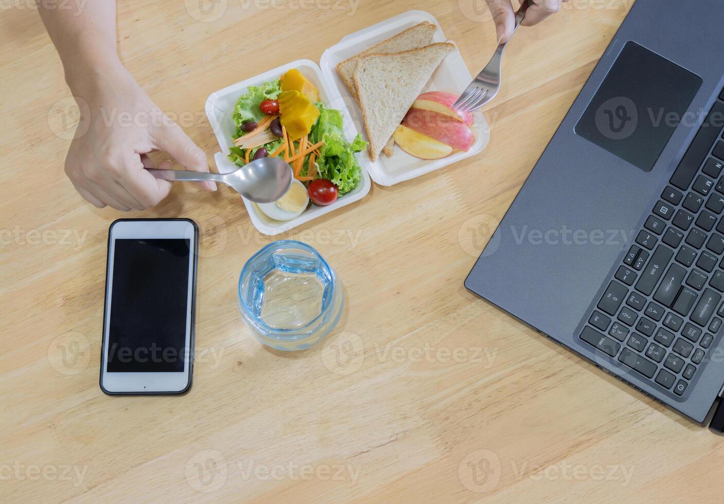 Woman eating tasty healthy salad on white wooden table with laptop and mobile phone. photo
