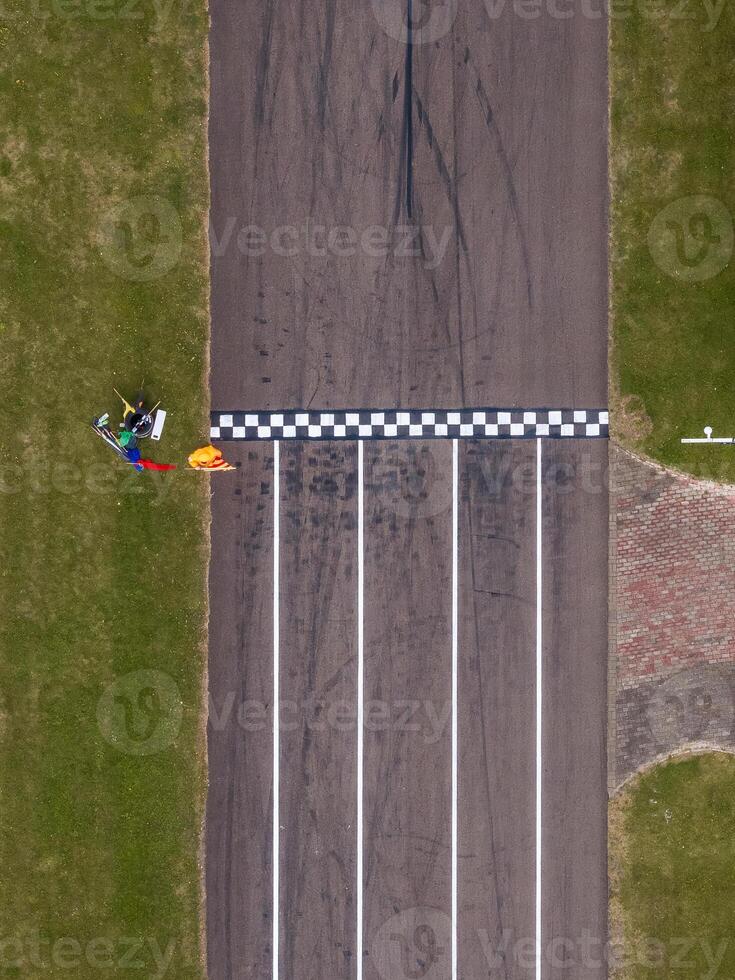 An aerial view of a race track featuring a checkered finish line, bordered by grass, with tire marks, equipment, and a person in an orange outfit visible. photo