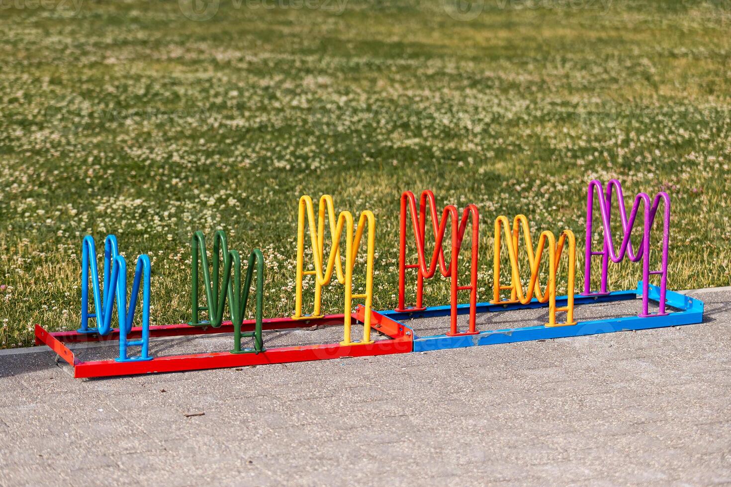 A vibrant bike rack with wavy loops in blue, green, yellow, red, and purple stands on a paved surface beside grass with small white flowers. photo