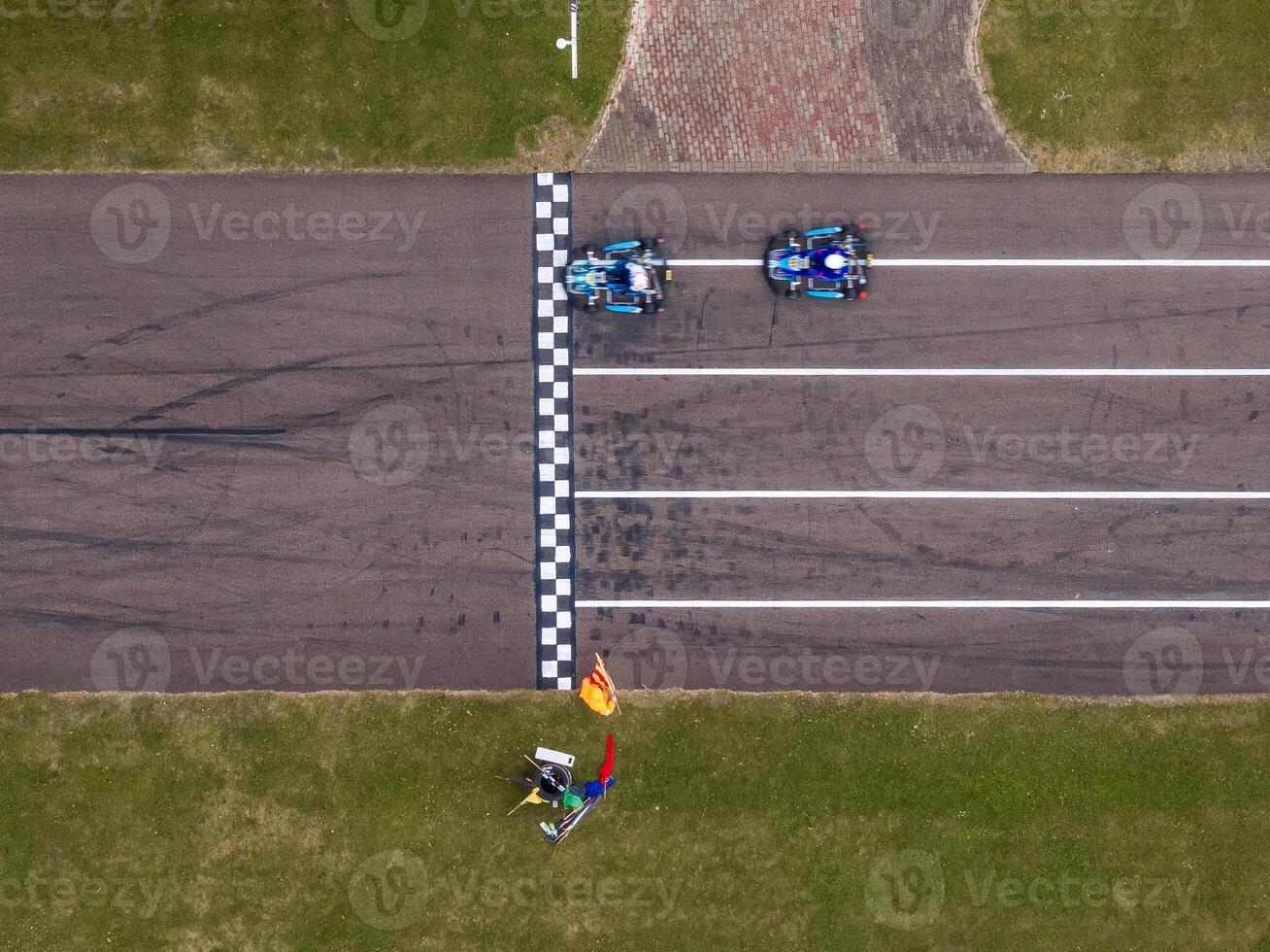 Two go karts cross a checkered finish line on a paved track, with a race official holding flags nearby. Tire marks and a brick walkway are visible. photo