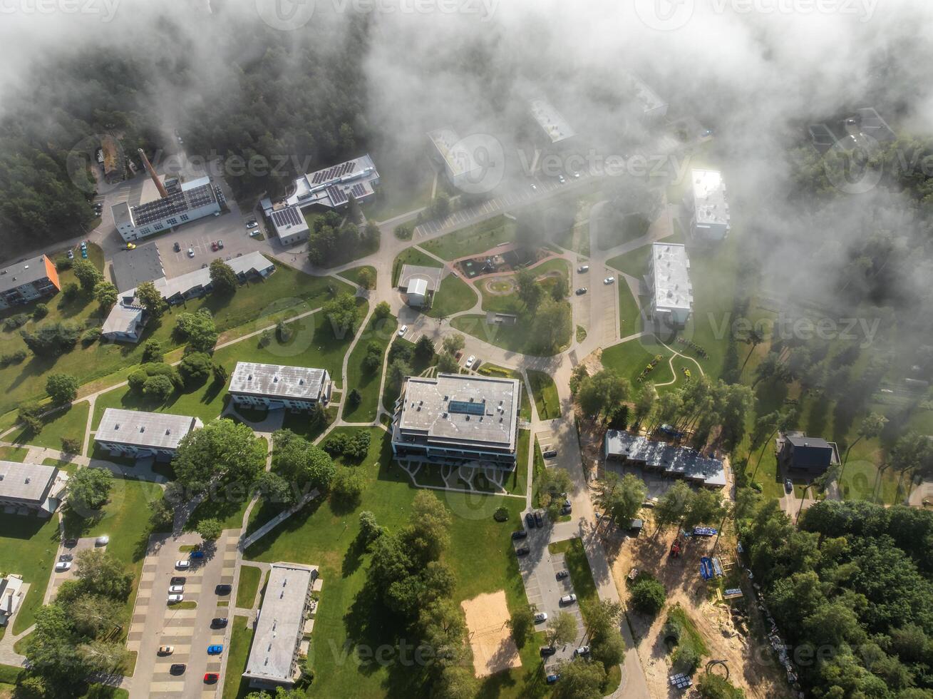 Cluster of modern buildings surrounded by trees and open green spaces, with pathways, parking lots, and light fog partially obscuring the scene. photo