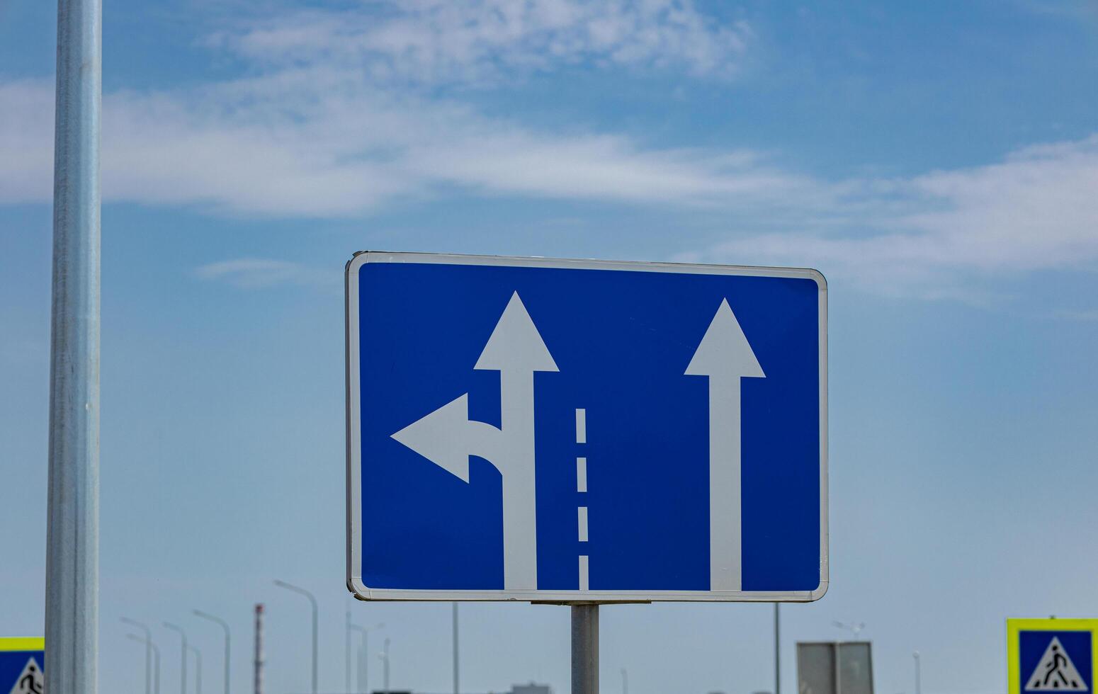 A traffic sign shows arrows indicating left and straight directions for vehicles on a road with a blue sky above photo