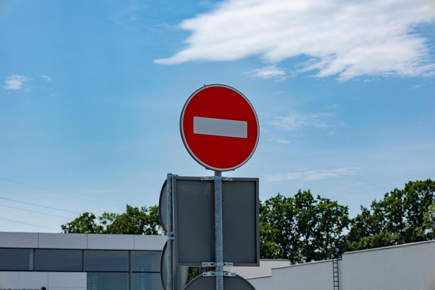 A bright red no entry sign is visible against a clear blue sky, surrounded by greenery and a modern building in the background photo
