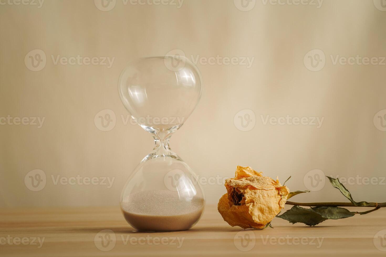 A simple hourglass on a wooden table with dry rose a beige backdrop, symbolizing time flow, aging, and minimalistic living. Represents timeless themes in a creative presentation for stylish concepts. photo