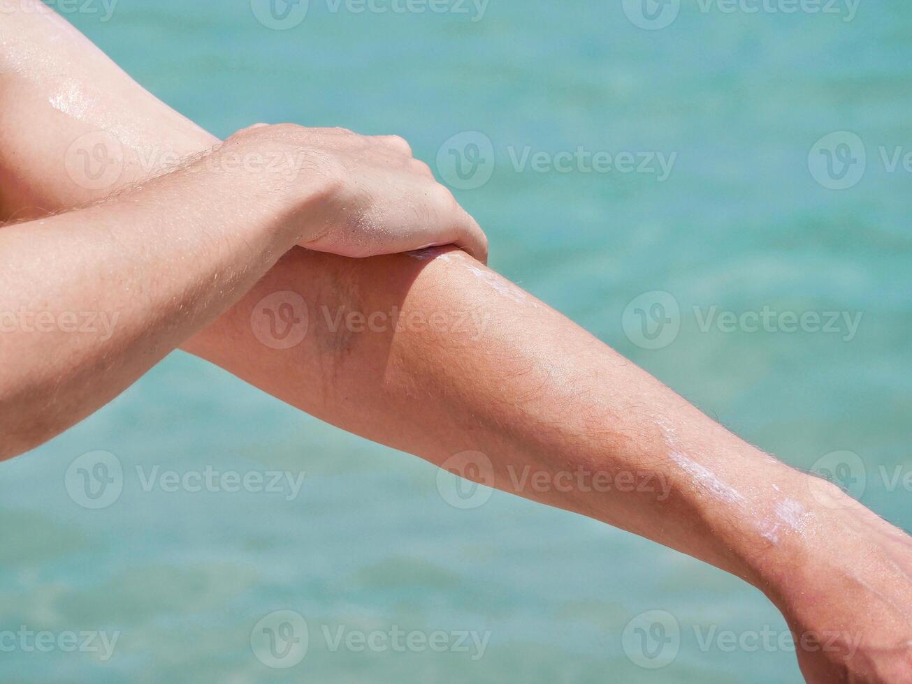 A man on the beach applies sunscreen lotion to his hands at the background of sea water. Skincare, sun protection and beach holidays concept. photo