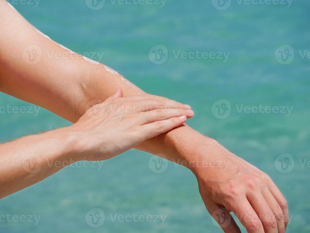 A man on the beach applies sunscreen lotion to his hands at the background of sea water. Skincare, sun protection and beach holidays concept photo