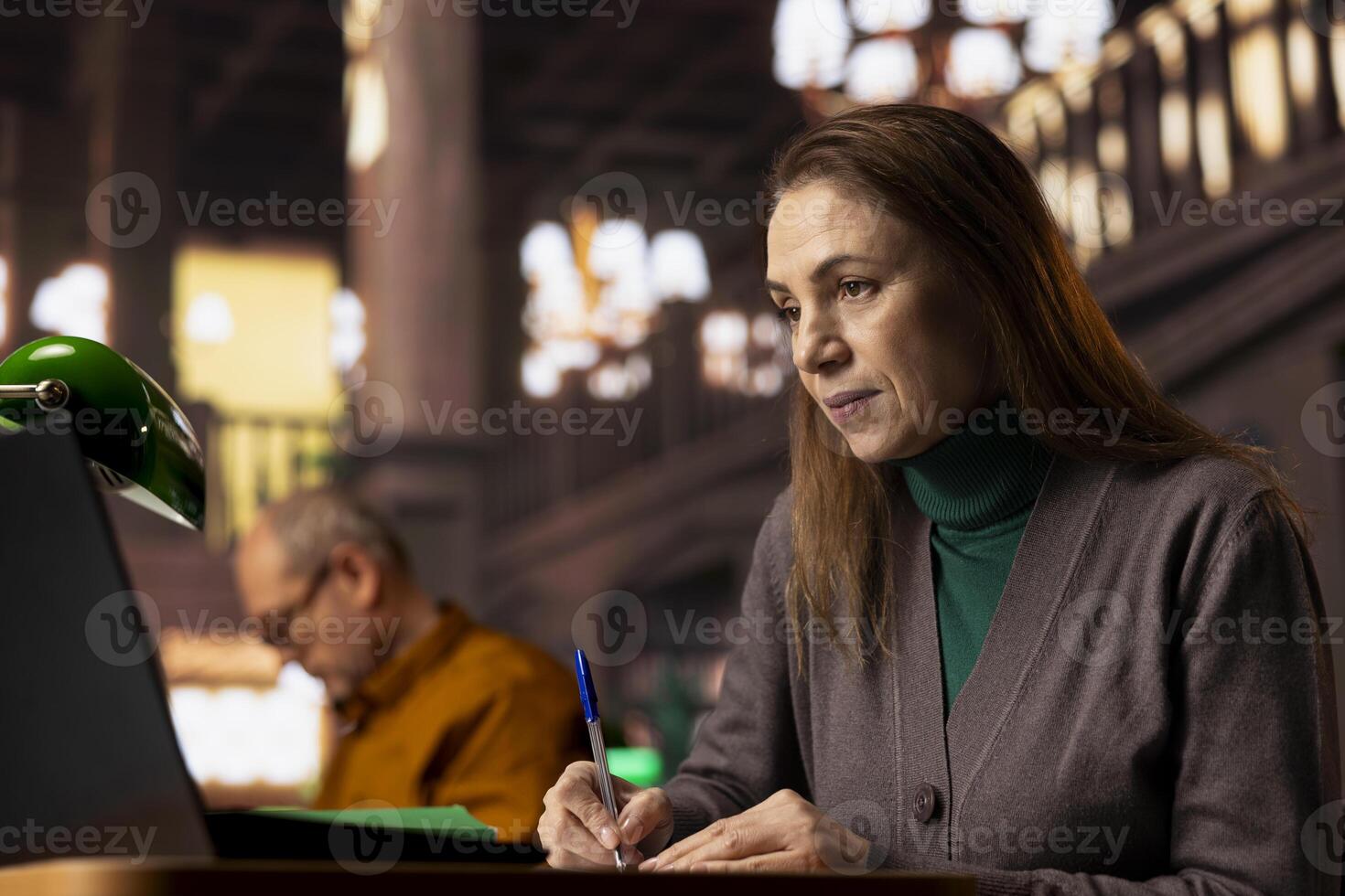 Elderly academic woman prepares documents for her university coursework, transcribing from literature and textbooks. Student focused on research and education in a quiet public library. photo