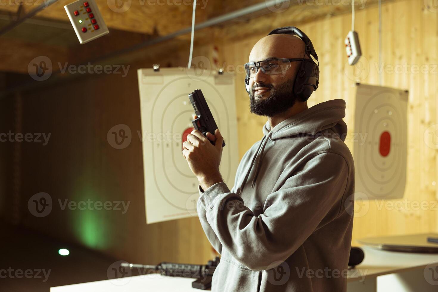 Arab firing range customer holding firearm enjoying recreational indoor shooting experience. Middle Eastern man wearing protective gear practicing with weapons in safe, welcoming environment photo