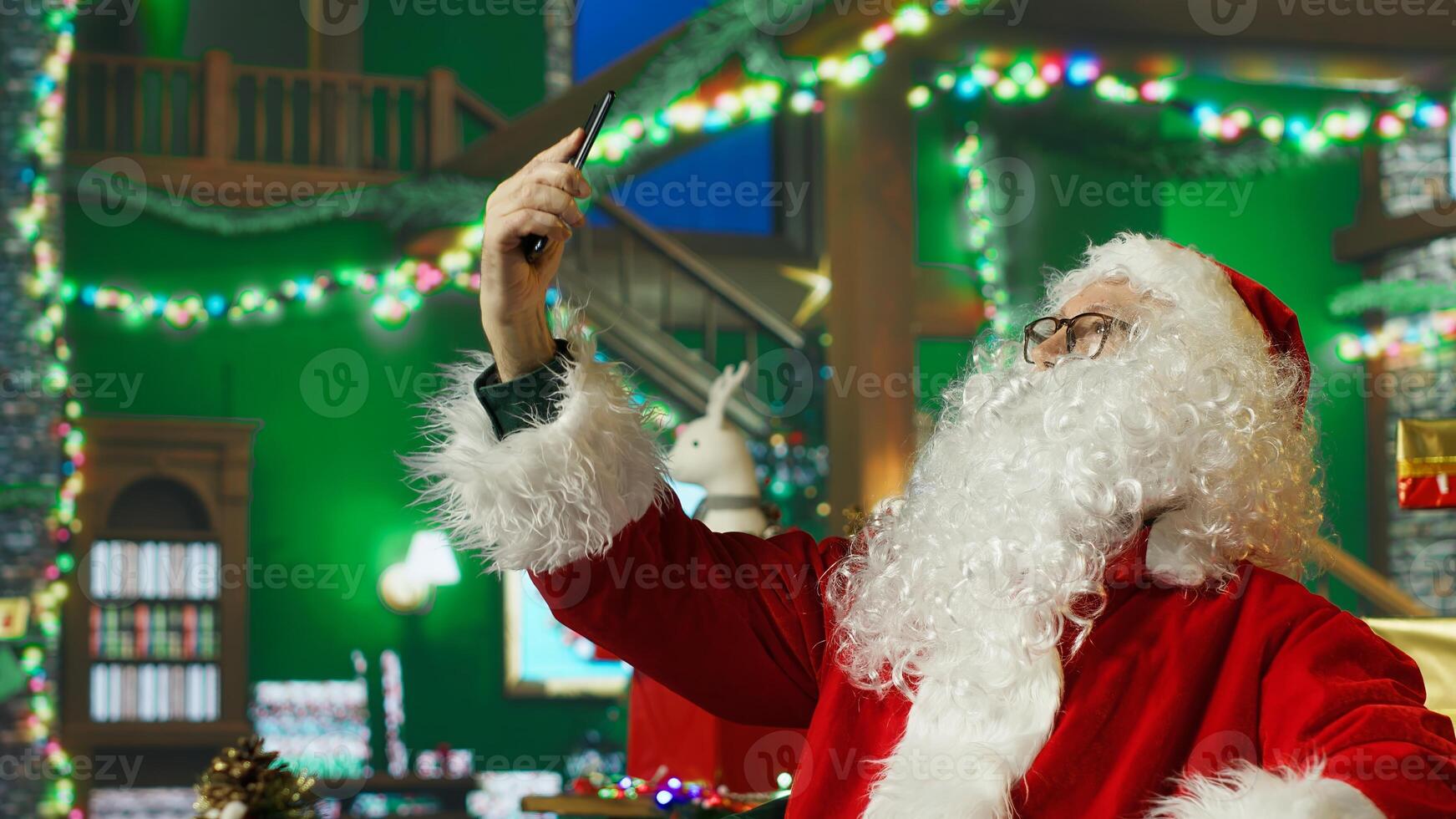Professional Santa Claus taking phone selfie photos on Christmas set with large chairs, piles of presents, backdrops, stockings, fake fireplace, ornaments and other holiday props, camera A