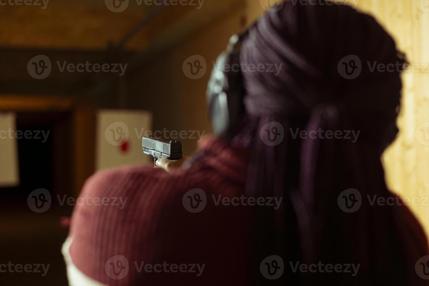 Woman in firing range doing shooting activity with pistol, wearing earmuffs to protect hearing. Person in shooting gallery training with firearm, using safety gear to prevent damage from gunshots photo