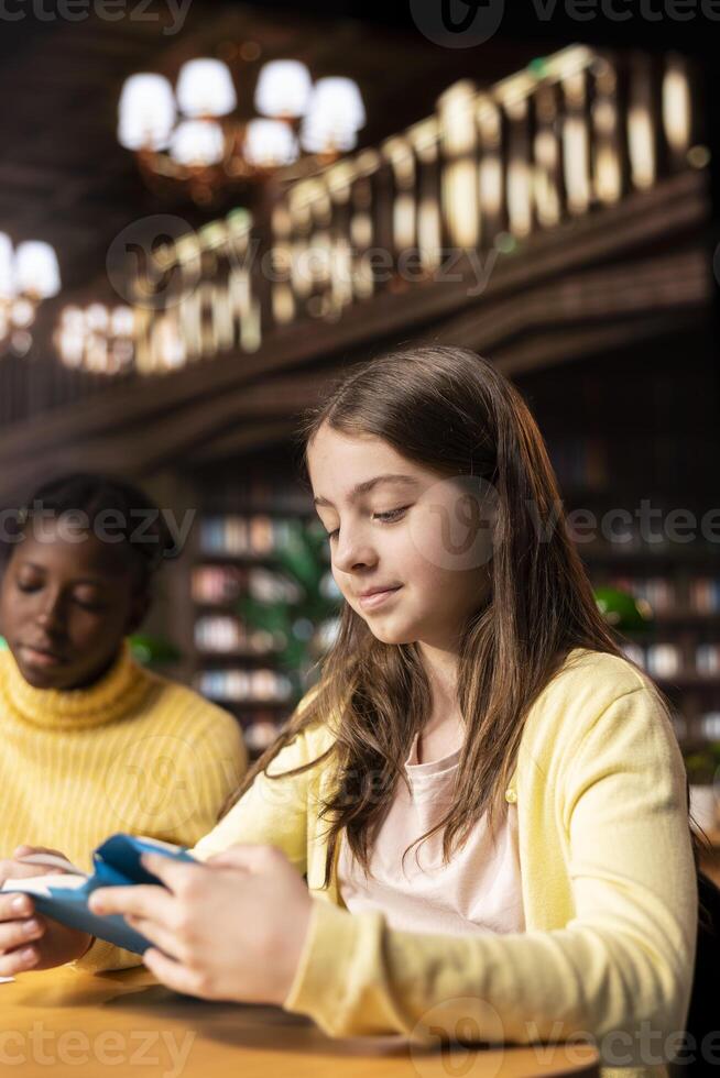 Two preteens girls reading books in the library to focus on mandatory literature and gaining knowledge for their academic curriculum during study time. Young scholars focusing on lecture tasks. photo