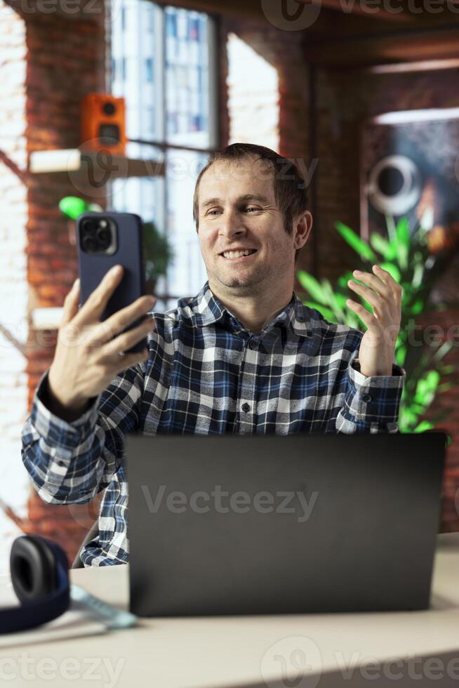 Joyous freelancer in home office sitting at desk in front of laptop, greeting colleagues in videocall. Cheerful employee in teleconference meeting with managers, answering inquiries photo