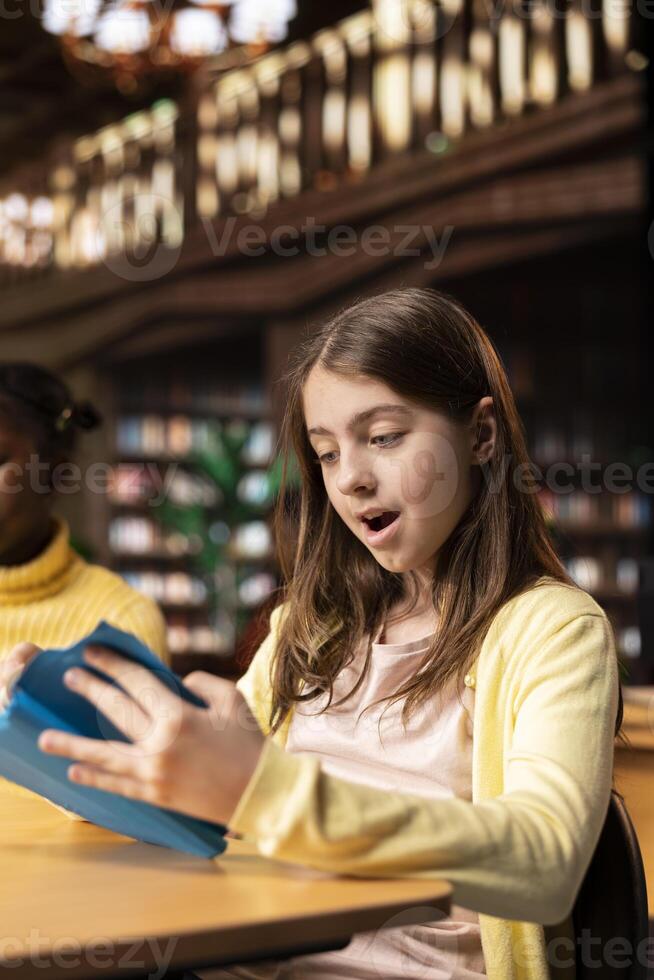 Surprised teenage girl enjoying mandatory literature book in the library, feeling amazed by the lecture story. Schoolgirl reading literary work from the annual school curriculum, academic success. photo