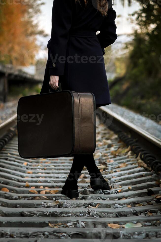 Woman in Black coat with Vintage suitcase walks along railway path in autumn, surrounded by vibrant fall leaves, capturing essence of travel and exploration. photo