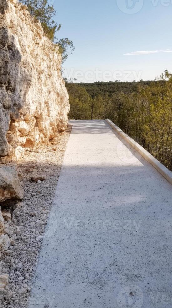 Sun-dappled pathway alongside a rugged rock face, leading into a serene natural landscape with lush green trees under a clear sky. photo